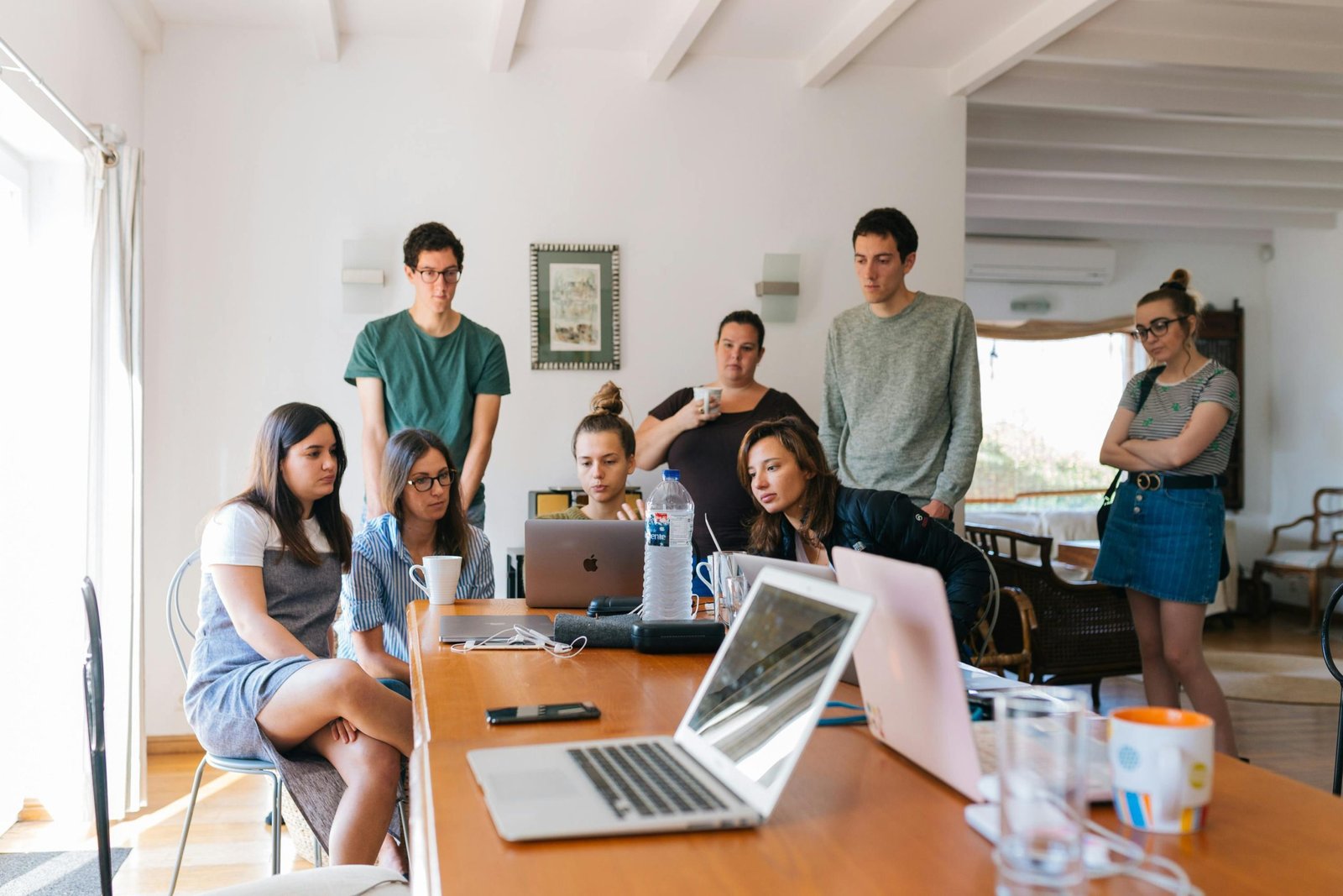 A group of people sitting and standing around a table, engaging with laptops and a smartphone. The well-lit room, designed by Irina Stoica designer interior, features white walls and elegant wooden furniture, blending functionality with modern architecture.